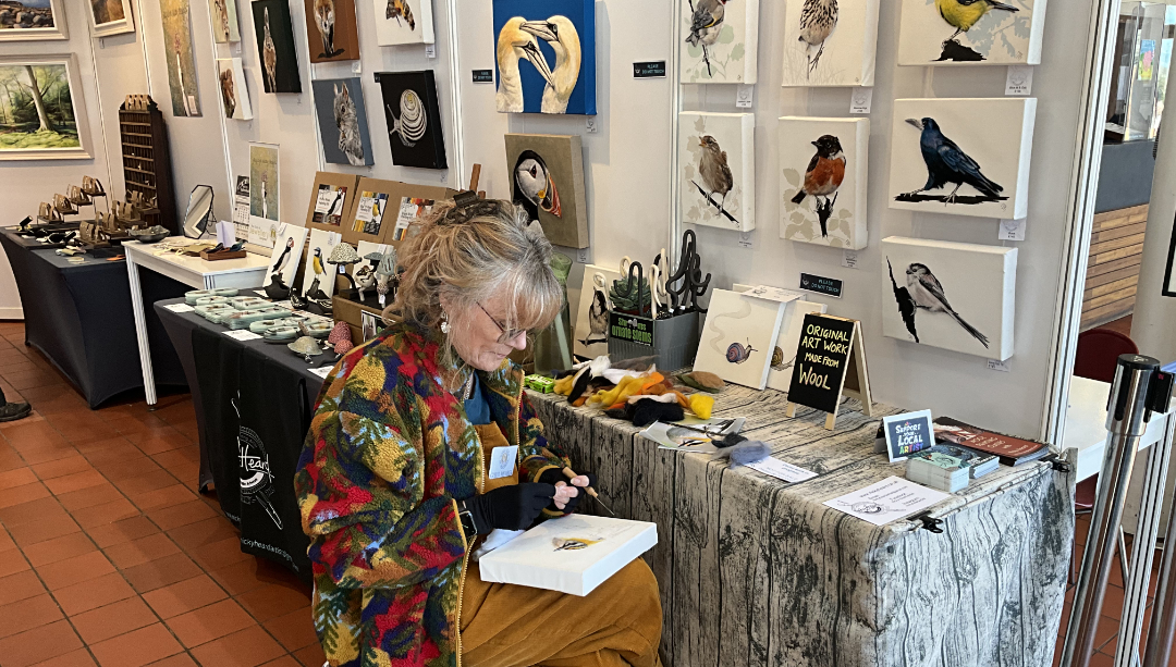 Nicky Heard seated with a canvas on her lap and demonstrating needle felting with her stand and other artwork in the background.